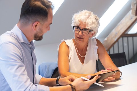 Eine Frau mit weissen Haaren und Brille bespricht mit einem Mann in einem hellen Büro Inhalte auf einem Tablet.