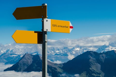 Wegweiser mit der Aufschrift «100% erneuerbar» vor schneebedeckten Bergen im Hintergrund.