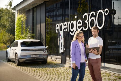 Une femme et un homme se tiennent devant le bâtiment d’Energie 360° et regardent ensemble une tablette, avec en arrière-plan une voiture électrique en train de se recharger à une borne.