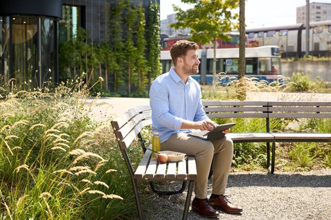 Une personne est assise sur un banc devant l'entrée principale et travaille sur sa tablette. Un tramway passe.