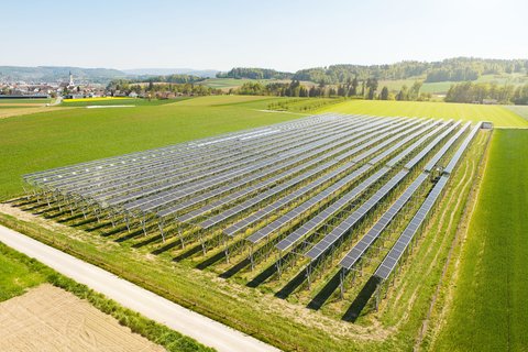 Installation agri-photovoltaïque avec de longues rangées de panneaux solaires au-dessus d’une surface agricole verdoyante, entourée de champs et de collines.