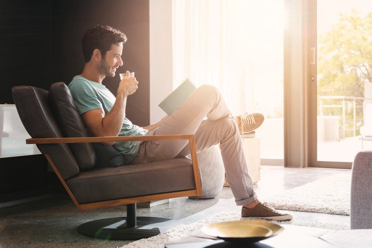 Mann sitzt im Wohnzimmer auf einem Stuhl, liest ein Buch und trinkt Kaffee