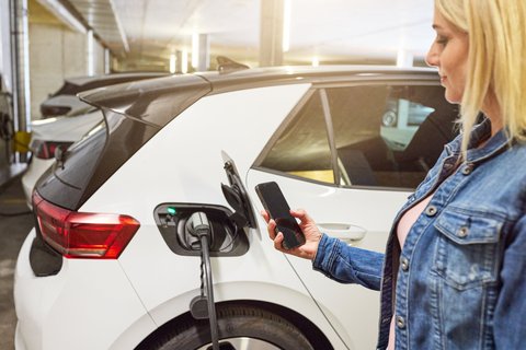 Eine Frau steht direkt neben ihrem Elektroauto und betrachtet auf ihrem Handy Informationen zu easycharge.
