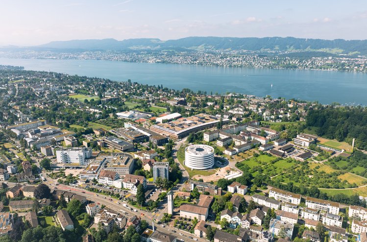 Luftaufnahme eines Quartiers mit mehreren Gebäuden, Grünflächen und rundem Neubau; im Hintergrund liegen See, Siedlungen und Hügelzüge.