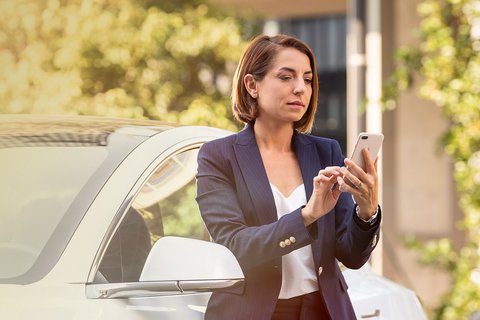 Une femme se tient juste à côté de sa voiture électrique et regarde sur son téléphone portable des informations concernant charge@destination