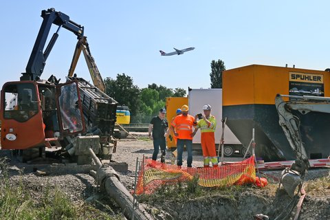 Baustelle neben dem Flughafen Zürich mit Bauarbeitern und Flugzeug im Hintergrund.