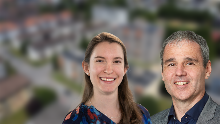 Zwei Personen besprechen in einem Büro Architekturpläne und Visualisierungen für ein Bauprojekt; eine Person zeigt mit einem Stift auf einen aufgehängten Plan.