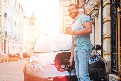Un homme se tient devant une voiture électrique. Dans une main, il tient un câble de recharge et dans l’autre, un gobelet en carton