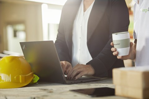 Une femme devant son ordinateur et une personne à côté d’elle avec un gobelet de café à la main